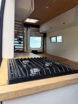 Inside an RV rental: a built-in three-burner gas stove on a wooden counter with a rustic van conversion interior and windows.