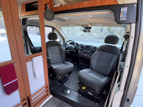 RV rental: Camper van interior. Front view of cockpit with driver/passenger seats, dashboard, and the road visible through the windshield.