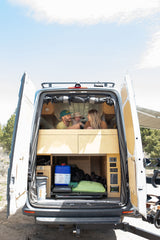 Family enjoying their RV rental in a camper van. Open rear doors reveal a cozy custom interior with storage below.