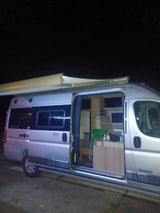 Night shot of a silver camper van, interior visible with a dining area & kitchen. An extended awning suggests camping. Great rv rental.