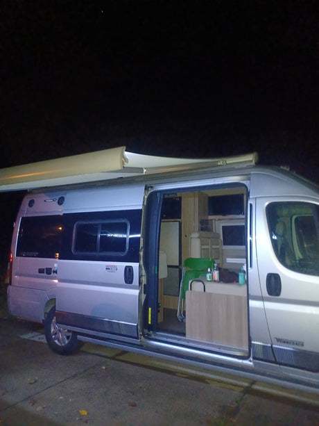 Night shot of a silver camper van, interior visible with a dining area & kitchen. An extended awning suggests camping. Great rv rental.