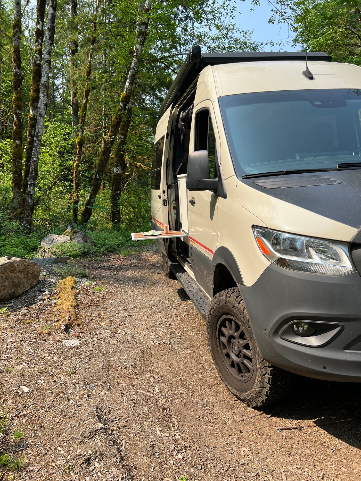 RV rental camper van parked in a forest. Side door open, revealing a pull-out table with a drink and plate. — photo 8