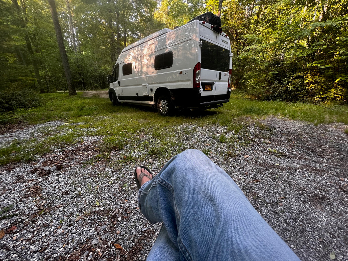 White Winnebago Solis rv rental van at a sunlit campsite among trees. Person's legs visible in foreground.