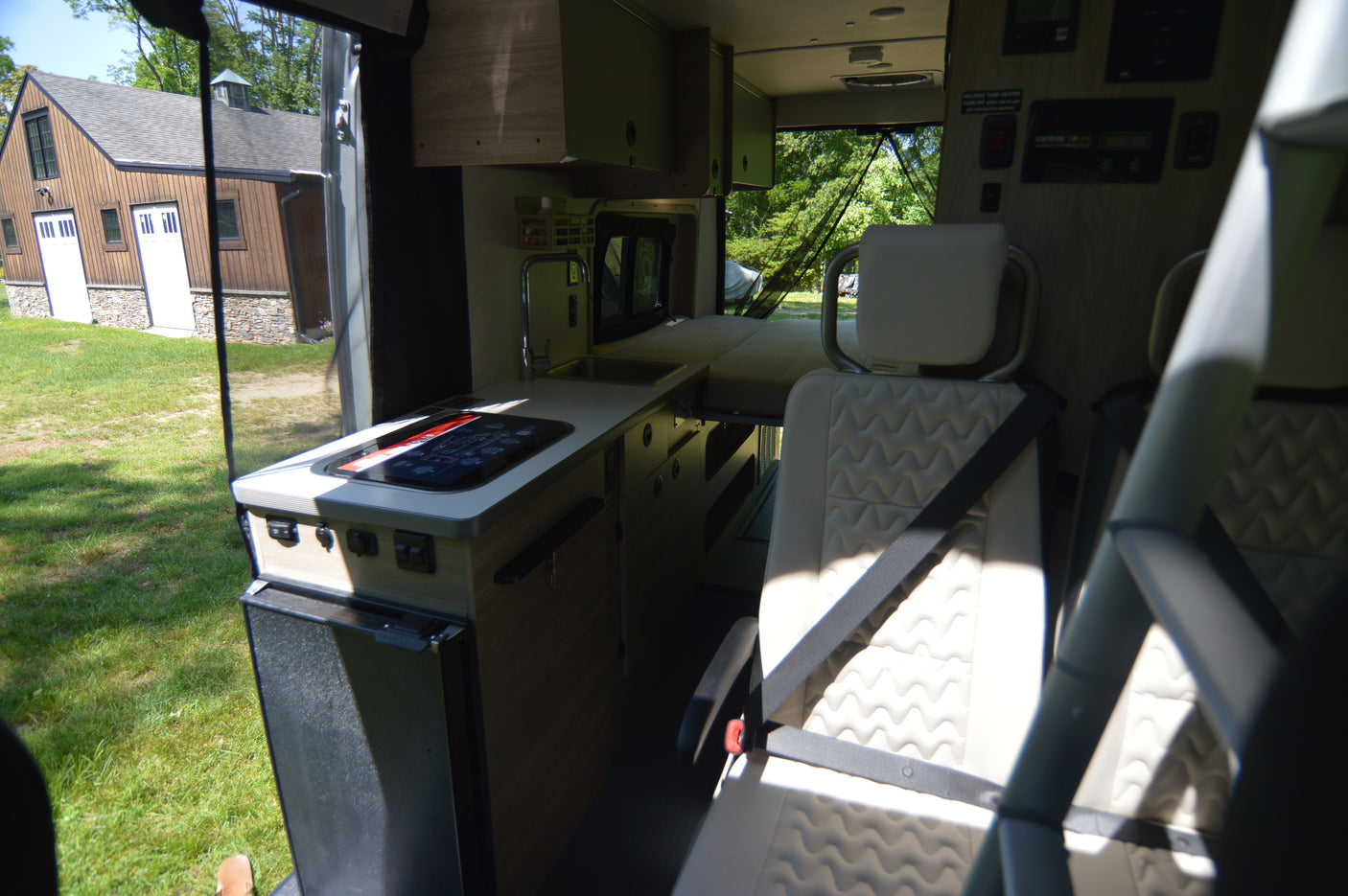 RV rental camper van interior: modern kitchen with cooktop, sink, and fridge; passenger seating; bed area. Barn view outside.