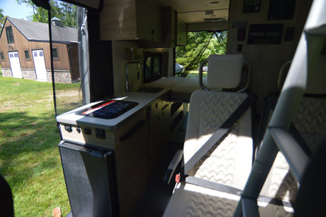 RV rental camper van interior: modern kitchen with cooktop, sink, and fridge; passenger seating; bed area. Barn view outside.