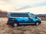 Custom-painted RV rental van featuring blue mountains and trees, parked in a grassy field under a cloudy sky.