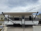 A white and gray Open Range travel trailer rv rental, with an extended awning, parked outside under a cloudy sky.