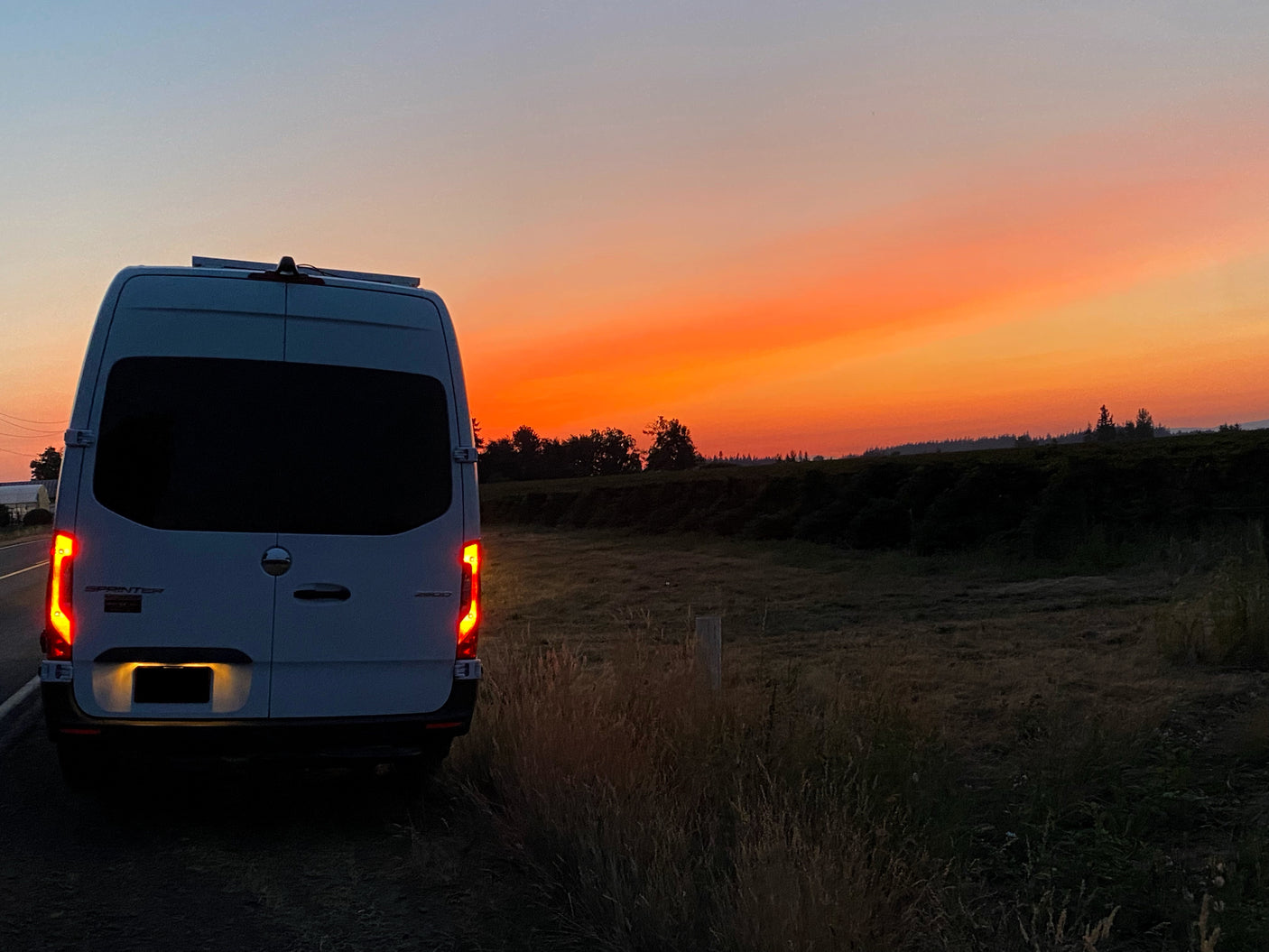 White van, likely an RV rental, parked on a rural road. Its taillights glow against a vibrant orange and red sunset sky.