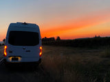White van, likely an RV rental, parked on a rural road. Its taillights glow against a vibrant orange and red sunset sky.