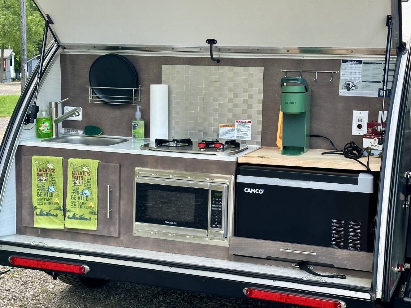  and an outdoor mat in a wooded campsite.|This rv rental features a compact outdoor kitchen with a sink — photo 5