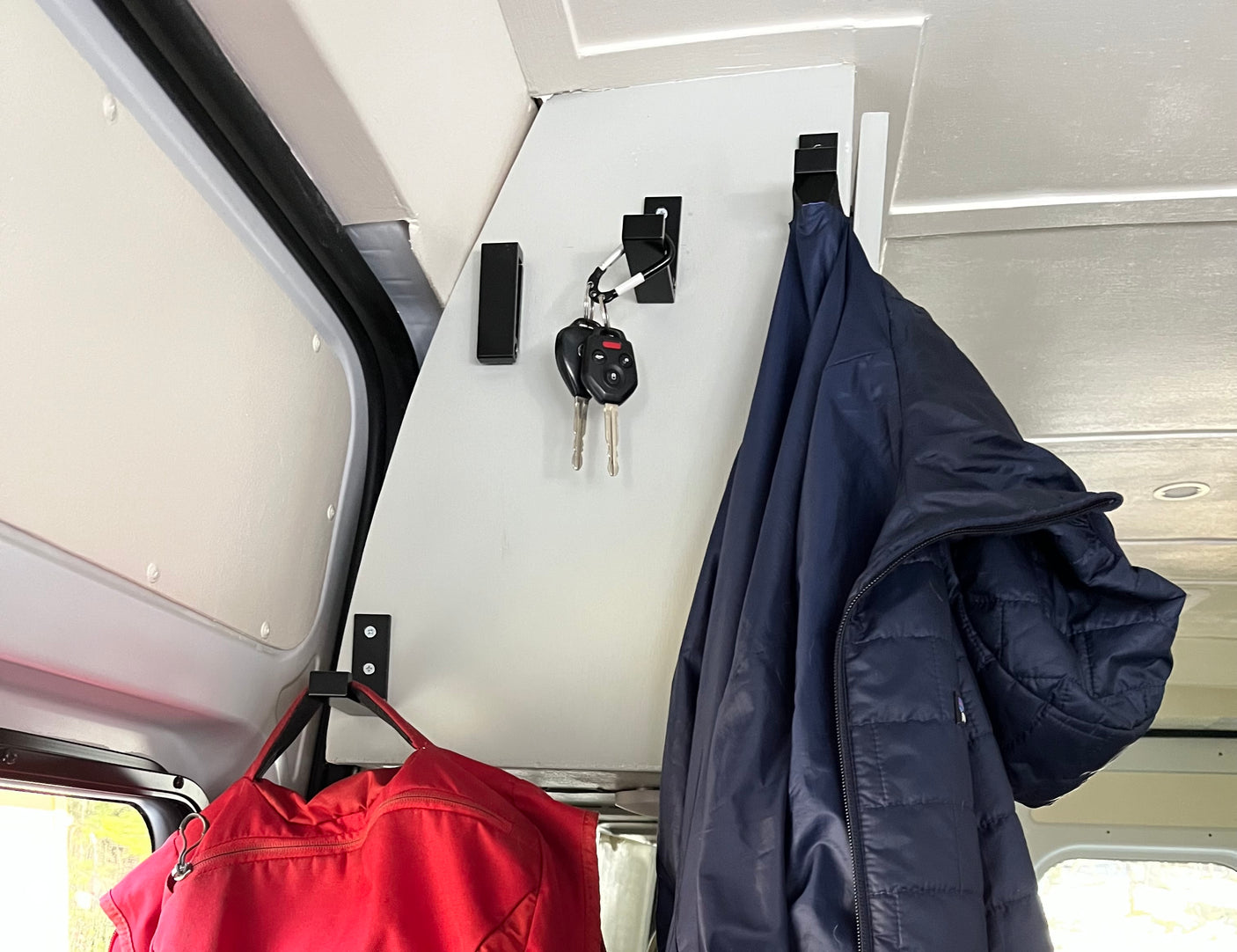 Interior of an RV rental with black hooks holding keys, a blue jacket, and a red bag for organized storage.