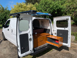 White camper van with side/rear doors open, revealing bed, cabinetry, and roof-mounted water tank/surfboards. Perfect for rv rental.