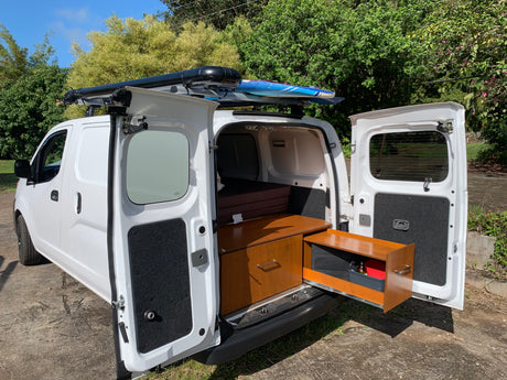 White camper van with side/rear doors open, revealing bed, cabinetry, and roof-mounted water tank/surfboards. Perfect for rv rental.