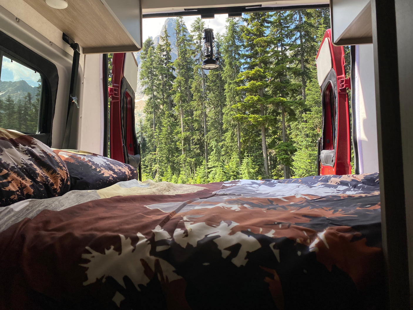 RV rental bed offers serene forest and mountain views from open back doors. A cozy spot in nature.