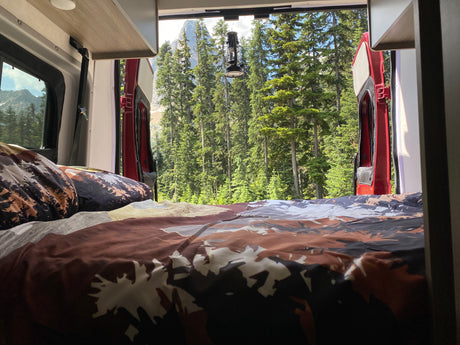 RV rental bed offers serene forest and mountain views from open back doors. A cozy spot in nature.
