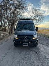 Adventure-ready black Mercedes Sprinter van, an RV rental, on a cracked road. Bare trees left, mountains right, sunset sky.