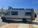 Side view of a light grey custom camper van with off-road tires, roof rack, and ladder. Perfect for an rv rental trip.