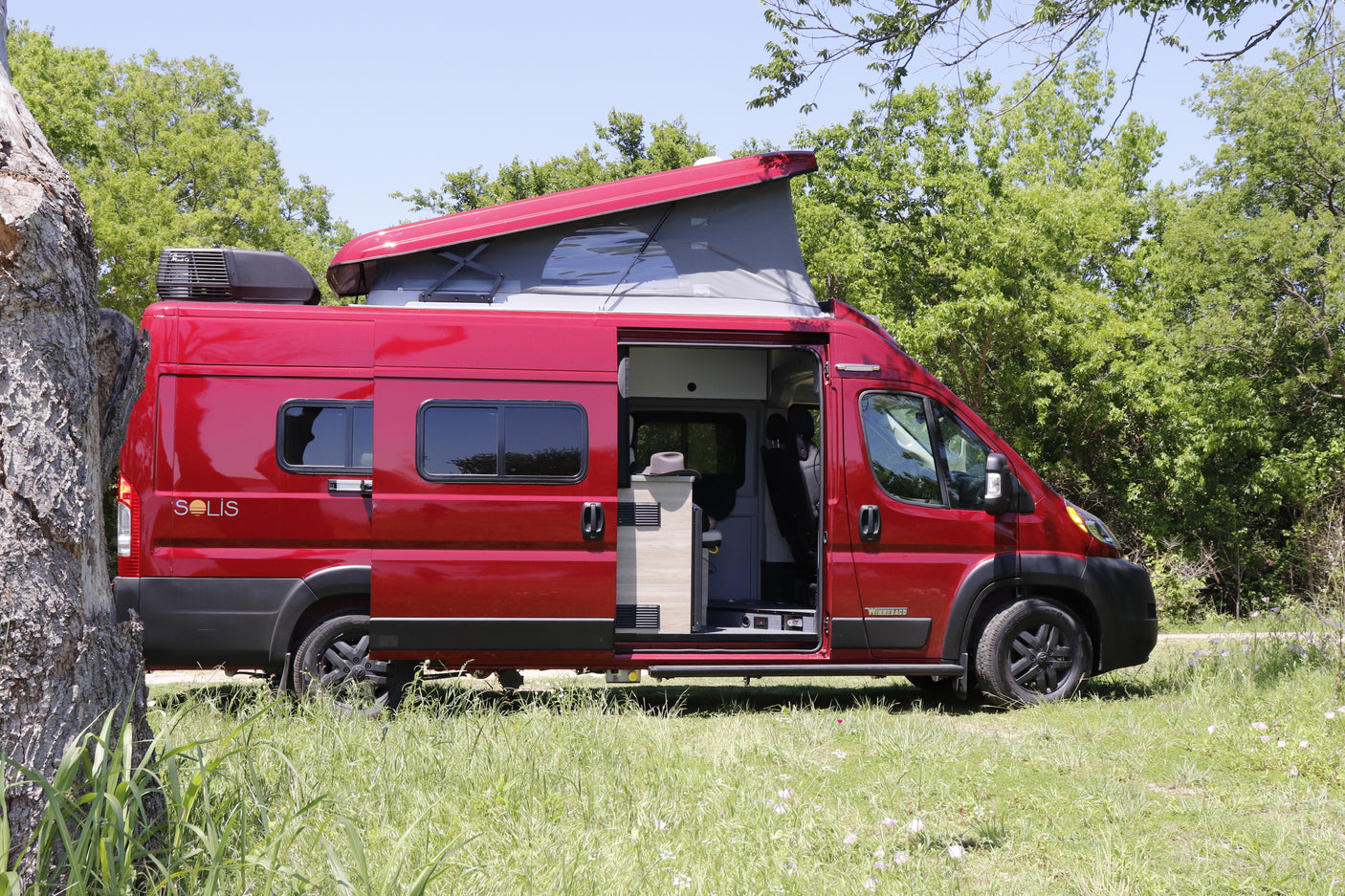 A red Winnebago Solis pop-top camper van (RV) parked in a grassy, treed area. Book your rv rental getaway.