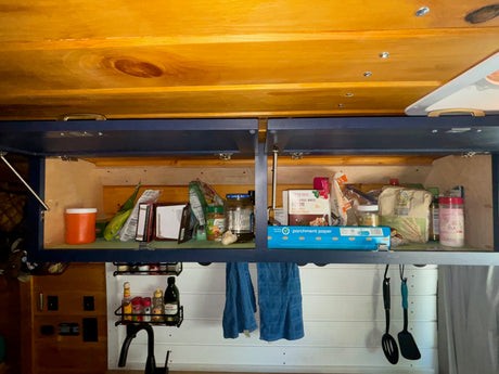 RV rental kitchen: open overhead cabinets reveal pantry essentials like food, parchment paper, spices, and utensils.